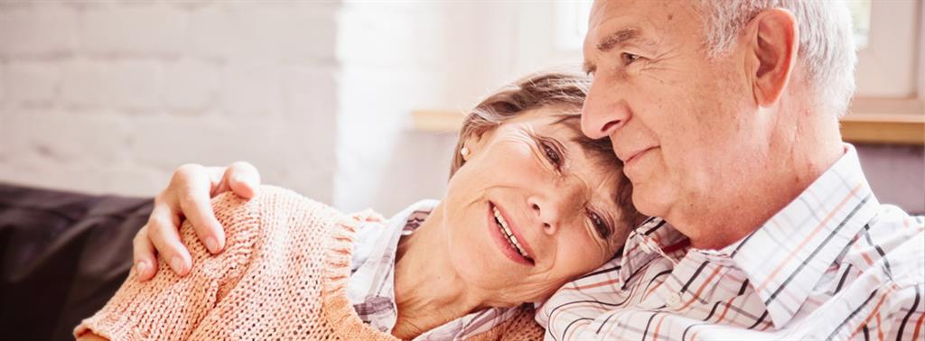 enior couple sitting down on a couch with husband’s arm wrapped around wife as she leans towards him content.